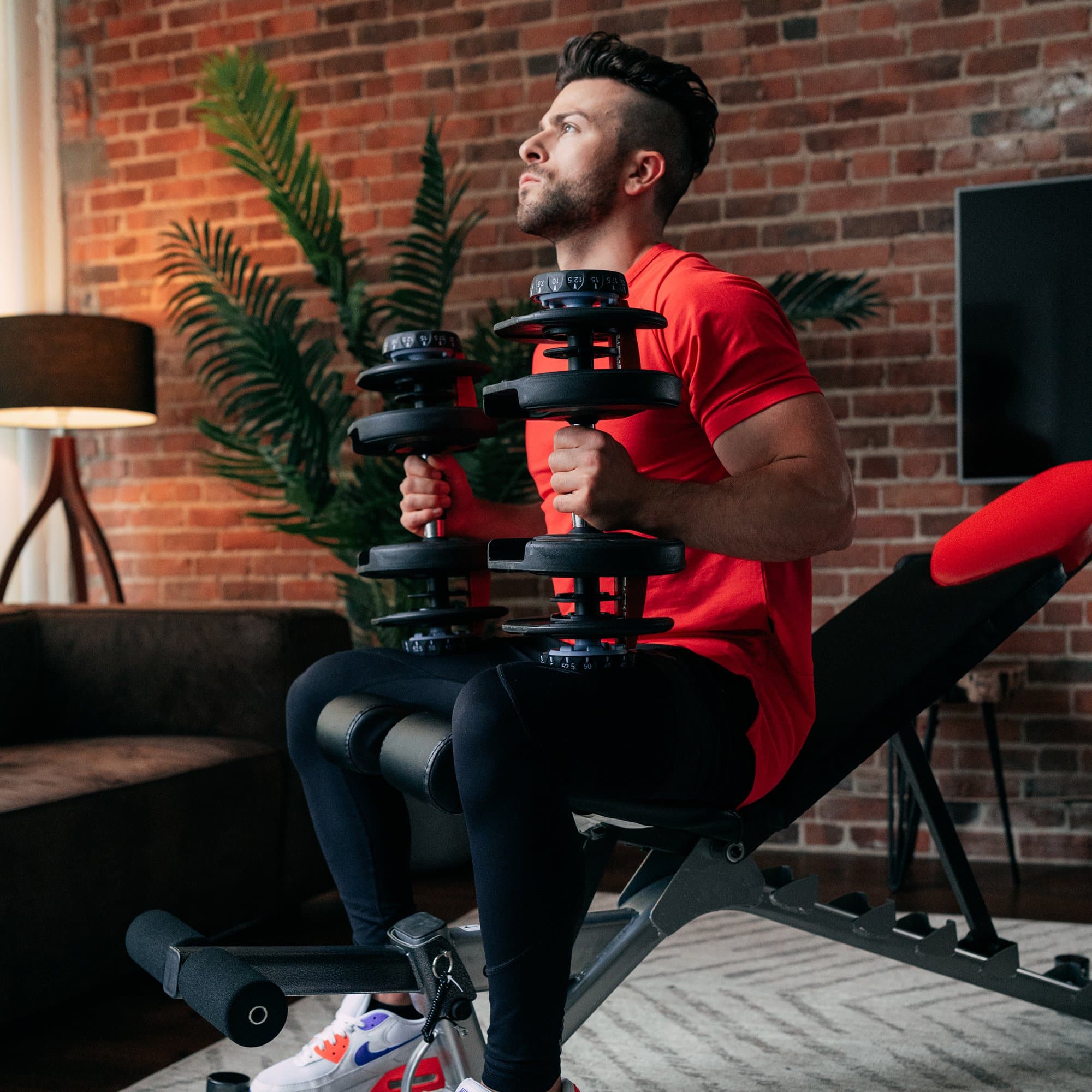 Man in red shirt lifting adjustable dumbbells on a bench in a brick-walled room.